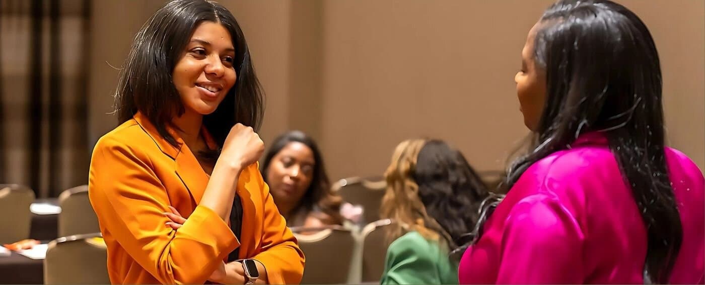 Jacqueline in an orange blazer, smiling during a conversation at an event.