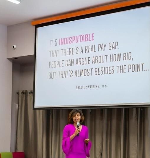 Jacqueline in a magenta dress on stage with a microphone, beneath a slide about the pay gap.